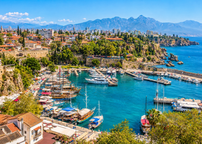 Barcos em uma marina diante de edifícios, vegetação e montanhas na costa do Mediterrâneo.